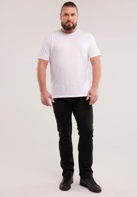 Mężczyzna z brodą i kucykiem stoi zwrócony przodem, ubrany w gładki biały t-shirt, czarne spodnie i czarne trampki na jednolitym tle.