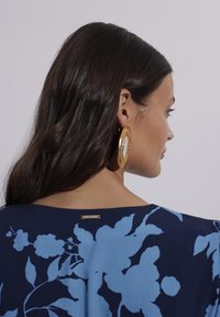 Granatowa bluzka w niebieski wzór kwiatowy, z dekoltem w serek i złotą metką na plecach. Modelka nosi duże, złote kolczyki w kształcie kół.