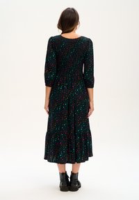 Žena s hnědými vlasy má na sobě černé midi šaty s barevnými malými leopardími skvrnami a černé kotníkové boty, otočená zády na bílém pozadí.