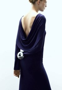 Długo rękawowa sukienka w głębokim purpurowym kolorze z drapowanym tyłem, z metalicznym akcentem w postaci kuli. Gładki materiał z eleganckim, dopasowanym krojem.