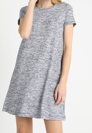 Kobieta ubrana w casualową sukienkę w krótkim rękawie, szarą z czarnymi cienkimi paskami, stojąca na jednolitym tle.