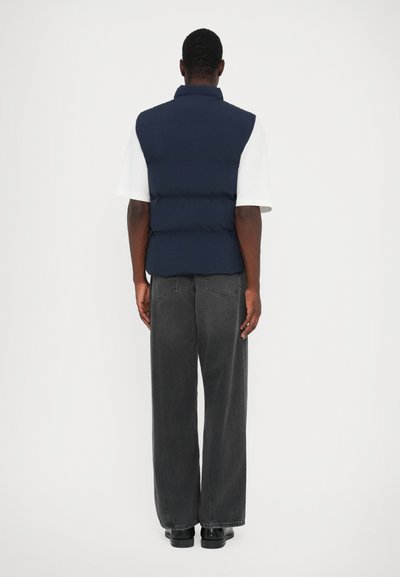 Vest puf navy blue cu guler înalt, design fără mâneci și secțiuni umplute, purtat peste o cămașă albă și blugi închiși la culoare.