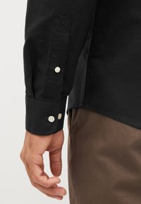 Cămașă neagră cu manșetă butonată și trei butoane albe, fabrică texturată și design cu manșetă ascuțită. Pantaloni maro parțial vizibili.