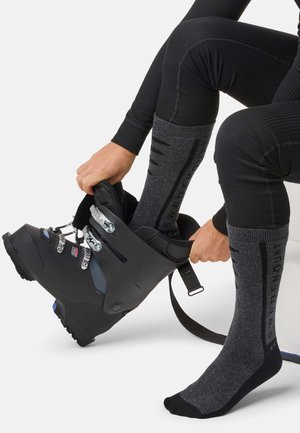 Une personne assise, vêtue de vêtements thermiques noirs, enfile une chaussure de ski noire par-dessus une chaussette de ski grise.