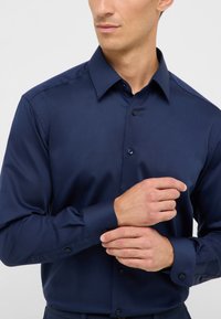 Camicia a maniche lunghe blu navy realizzata in tessuto morbido, con colletto a punta, bottoni frontali e polsini abbottonati. Presenta un design aderente.