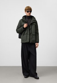 Zielona kurtka puchowa z kapturem i wieloma kieszeniami, noszona na czarnym golfie; zestawiona z szerokimi czarnymi spodniami i ciemnymi butami.