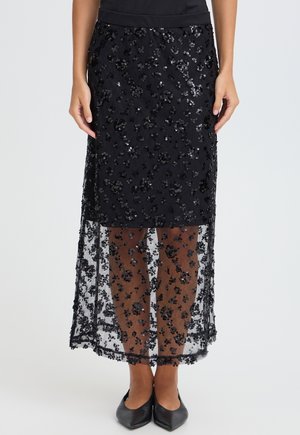 Jupe noire à sequins avec une couche intérieure solide se terminant au-dessus du genou et une superposition transparente ornée s'étendant jusqu'au milieu du mollet, portée avec des ballerines noires pointues.