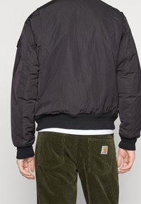 Svart vadderad bomberjacka med ribbade muddar och nederkant, bärs över gröna kordbyxor med ett etikett på bakfickan. Texturerad tyg synlig.