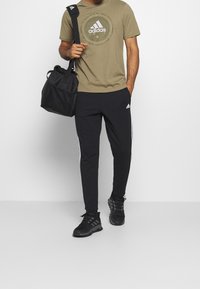 Sorte joggers med hvide sidestriber, olivengrøn Adidas t-shirt med logo, og sort sportstaske. Stylet med sorte træningssko.