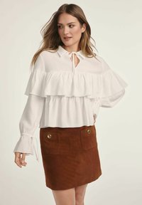 Biała bluzka z falbaniastymi warstwami i wiązanym dekoltem, zestawiona z brązową zamszową spódnicą z złotymi guzikami i dwiema przednimi kieszeniami.