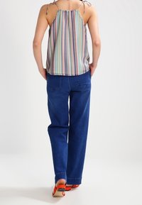 Person iført en stribet ærmeløs top med bindebånd, blå jeans og røde sandaler, stående med hænderne i baglommerne.