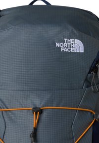 Dunkelgrauer The North Face Rucksack mit weißem Logo und orangefarbenem verstellbarem Bungee-Seil auf der Vorderseite.