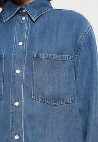 Πουκάμισο από τζιν ύφασμα, ανοιχτό μπλε, με μυτερό γιακά, κλείσιμο με κουμπιά και δύο τσέπες στο στήθος. Υφή υφάσματος με εμφανείς ραφές.