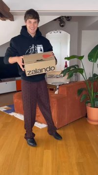 Kartonowe pudełko oznaczone napisem "zalando" trzymane przez osobę, z okrągłą czerwoną naklejką. W tle znajduje się roślina i pomarańczowa kanapa.