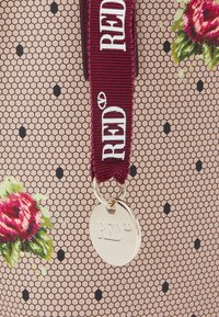 Sac à main à motifs avec un design en maille hexagonale rose agrémenté de pois noirs, souligné par des détails floraux et une bandoulière bordeaux avec une étiquette en métal.