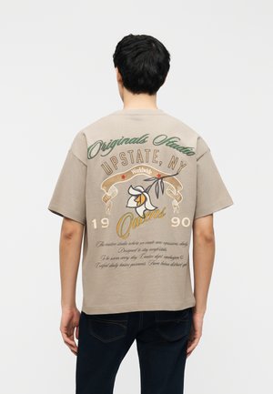 Muž nosiaci béžové tričko s kvetinovou grafikou a textom "Originals Studio Upstate, NY Queens 1990" vytlačeným na chrbte.