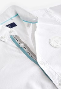 Valge nööbitav särk, mille krae sees on sinine ääris, Ted Bakeri bränditud nööbid ja ümmargune Ted Bakeri logoga plaastriga rinnal.