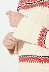 Maglione lavorato a maglia beige con polsini a costine, corpo texturizzato e un motivo geometrico rosso e nero intorno al colletto.