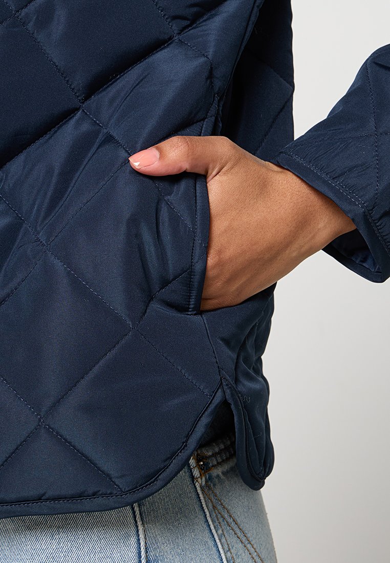 Chaqueta acolchada azul marino con superficie texturizada, un bolsillo lateral y un corte relajado con un dobladillo ligeramente curvado.