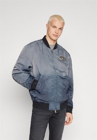 Veste bombardier dégradé bleu, avec fermeture éclair, poches latérales et poignets côtelés. Fabriquée en tissu léger avec un effet brillant.