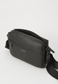 Black crossbody bag with textured leather, zip closure, adjustable strap, and subtle branding. Features a flat front pocket.