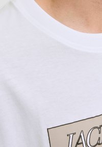 Camiseta de algodón blanca con cuello redondo, que presenta un acento gráfico beige con el texto "JAC." Textura suave y corte casual.
