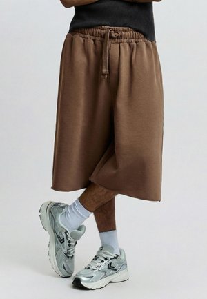 Henkilö, jolla on ruskeat polvenmittaiset shortsit, valkoiset puuvillasukat ja hopeanväriset urheilukengät, jalat ristiin yksinkertaisen taustan edessä.