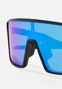Las grandes gafas de sol con montura negra presentan una lente degradada que va del azul al púrpura, con un diseño geométrico audaz y forma angular.