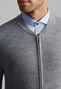 Un pull zippé gris avec un motif texturé et un col côtelé, superposé à une chemise à boutons bleu clair avec un bouton noir.