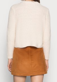 Cremefarbener Strickpullover mit kürzerem Schnitt und gerippter Struktur, kombiniert mit einem rostfarbenen Cord-Minirock mit vertikalen Streifen.