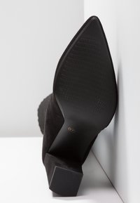 Bottine pour femme en daim noir avec bout pointu et talon carré, semelle montrant un motif de grip texturé, taille 37 indiquée sur la semelle.