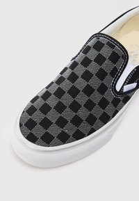 Fekete és szürke kockás slip-on sportcipő fehér gumitalppal és rugalmas oldalrésszel sima háttér előtt.
