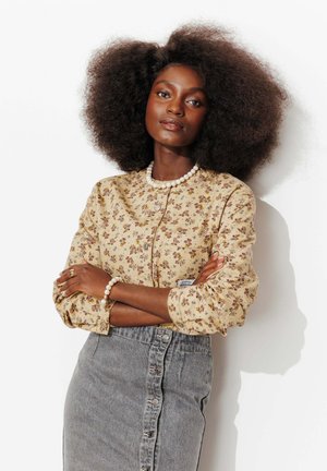 Femme confiante avec une grande coiffure afro naturelle, portant un chemisier beige à motifs floraux, une jupe grise boutonnée, ainsi qu'un collier et un bracelet en perles.