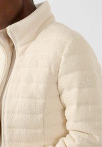 Chaqueta de forro polar beige claro con una superficie texturizada, cuello alto y un sutil detalle de branding en el hombro. Diseño acolchado visible.