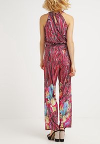 Vrouw in een mouwloze, met het rugpand gestrikte, rode jumpsuit met wijde pijpen en grote kleurrijke bloemenprints aan de onderkant.