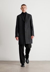 Cappotto di lana nera con colletto a revers, design monopetto e tre bottoni. Indossato sopra un dolcevita nero e abbinato a pantaloni neri.