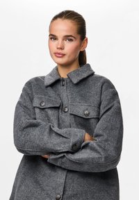 Γκρίζο, υφασμένο πουλόβερ από μαλλί με γιακά, κλείσιμο με κουμπιά και δύο τσέπες στο στήθος. Η εφαρμογή είναι χαλαρή με μακρύ μανίκι.