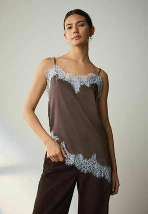 Young woman with dark hair wears a brown sleeveless top with light blue lace trim and dark pants, posing against a plain background.