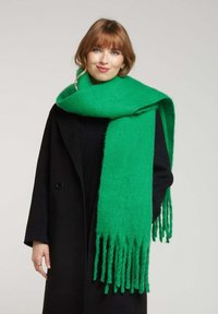 Écharpe verte à bords frangés en tissu doux, drapée de manière stylée autour du cou, avec une texture lisse et une couleur vive contrastant avec un manteau noir.