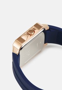 Rechteckige Uhr mit einem Gehäuse aus Roségold und einem navyblauen Silikonarmband. Verfügt über einen strukturierten Rücken und eine seitliche Krone zur Einstellung.