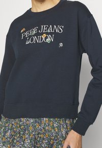 Felpa blu navy con la scritta "PEPE JEANS LONDON" ricamata, scollatura rotonda e polsini a coste. Abbinata a una gonna a fantasia floreale.