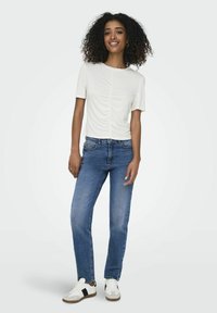 Top blanco de manga corta con detalle fruncido en la parte delantera, combinado con jeans de mezclilla azules y zapatillas blancas con acentos negros.