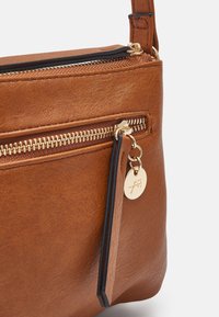 Borsa in pelle marrone con zip dorata e ciondolo rotondo dorato attaccato al cursore della zip, che mostra dettagliata texture e cuciture.