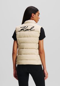 Beige Steppweste mit genähten Abschnitten und hohem Kragen. Auf der Rückseite befindet sich das schwarze "Karl"-Logo. Wird über einem schwarzen Shirt getragen.