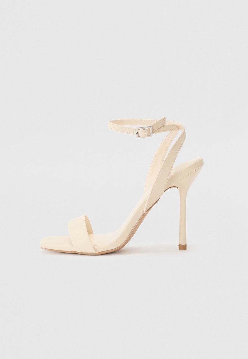 Beige høye hæler med en slank ankelrem og firkantet tå. Glatt syntetisk materiale, minimalistisk design, og stiletthæl.