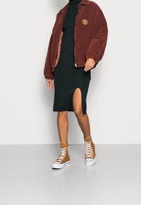 Braune Corduroy Bomberjacke, schwarzes Midi-Kleid mit seitlichem Schlitz und helle High-Top-Sneaker mit weißen Sohlen und Schnürsenkeln.