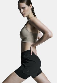 Beige sportieve crop top met textuurdesign, gecombineerd met zwarte shorts met een logo en een relaxte pasvorm. Model poseert in profiel.