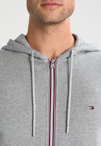 Grau melierte Zip-Hoodie aus weichem Material mit einem Frontreißverschluss, der ein rotes, weißes und blaues gestreiftes Design aufweist, sowie einem kleinen Logo auf der Brust.