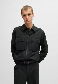 Zwart ritshemd van een textuurstof, met twee voorzakken en een omgeslagen kraag. Gecombineerd met bijpassende zwarte broek en een riem.