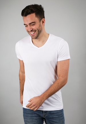 Glunderende man met kort donker haar, gekleed in een wit V-hals T-shirt en een blauwe jeans, staand tegen een effen grijze achtergrond.